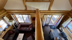 A wooden cabin interior with a loft bed, a sofa, a dining table, and large windows offering a view of the outdoors.