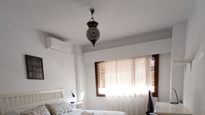 Een slaapkamer met een bed, een bureau, een stoel, een raam met jaloezieën en een hanglamp.