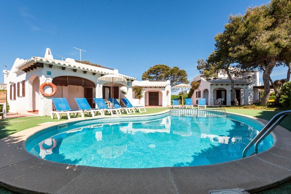 Una piscina con sillas de descanso y un flotador salvavidas, rodeada de edificios con arcos y un cielo azul despejado.