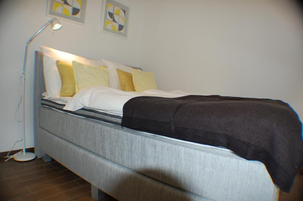 Uma cama de solteiro com colchão cinza, almofadas brancas e amarelas e um cobertor preto. Uma luminária de chão está do lado esquerdo da cama.