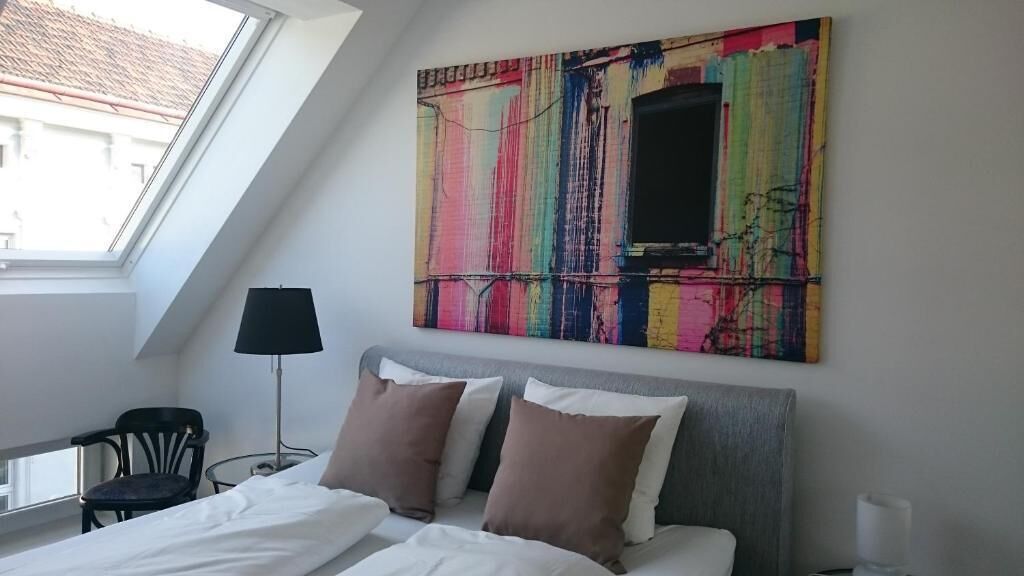 Quarto moderno com teto inclinado, uma grande pintura abstrata, cama com roupa de cama branca e marrom, abajur e uma cadeira.