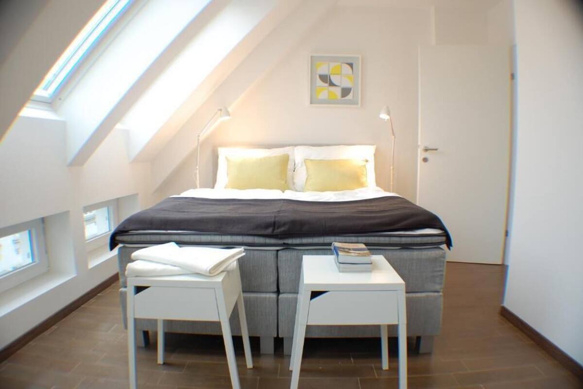 A modern bedroom with a sloped ceiling, a bed with a black and white duvet, two bedside tables, and a small bedside lamp.