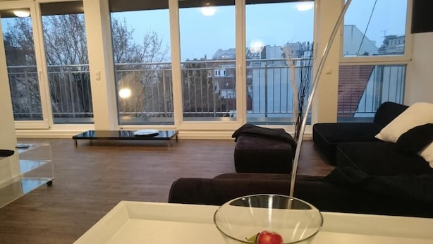 Un salon moderne avec une grande fenêtre, un canapé noir, une table basse blanche et un saladier en verre.