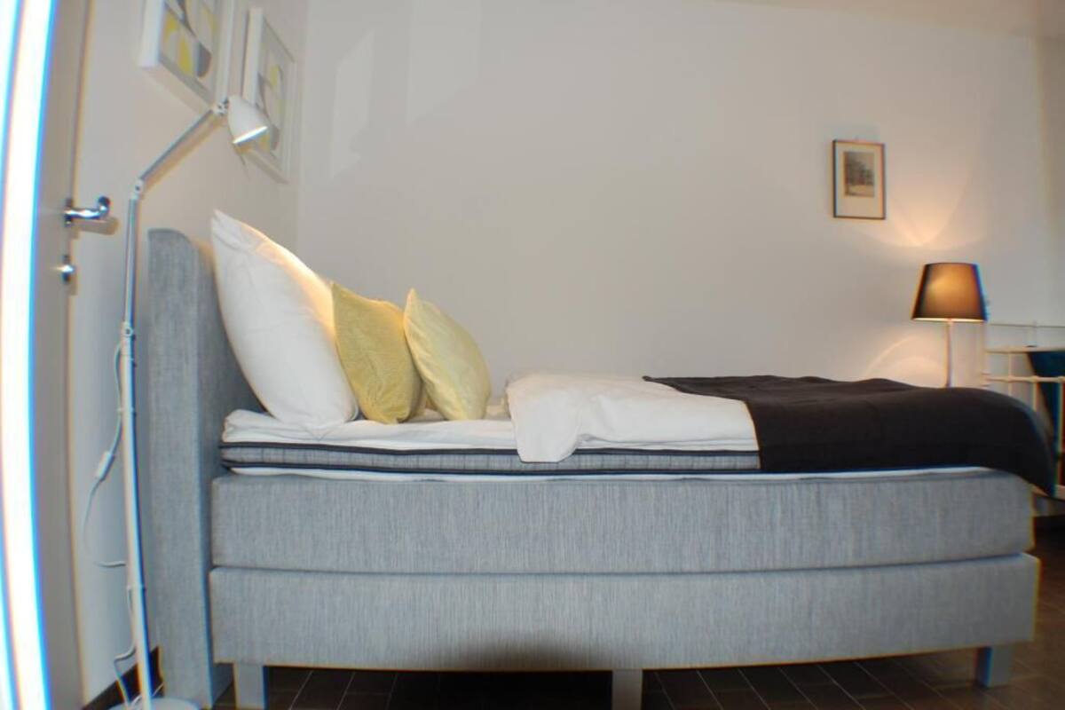 A modern bedroom with a grey sofa bed, white and yellow pillows, a black throw blanket, a bedside table with a lamp, and a framed picture on the wall.