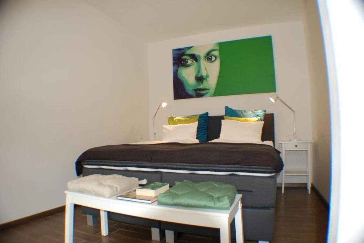 A bedroom with a bed, bedside tables, a bedside lamp, a pillow, a blanket, and a small table with books and a green cushion.