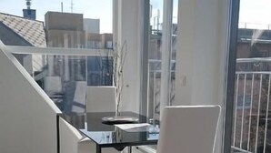 Un espace de repas moderne avec une table en verre et des chaises blanches, de grandes fenêtres offrant une vue sur le paysage urbain et un plancher en bois.