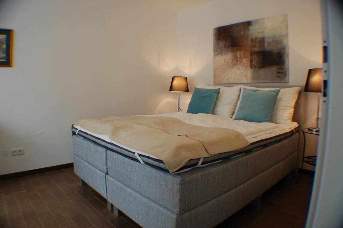 A neatly made bed with a beige cover, two pillows, and two bedside lamps.
