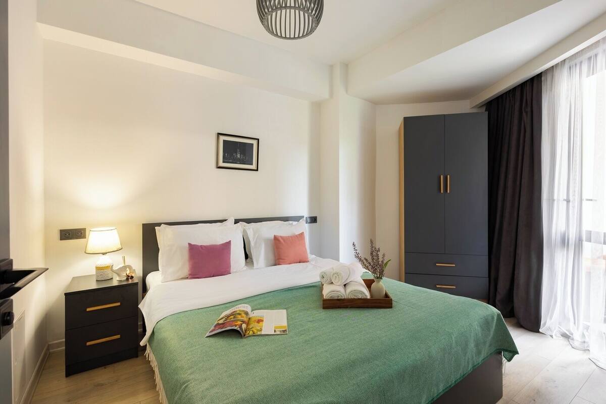 A hotel room with a bed, bedside table, lamp, wardrobe, and a window with curtains.