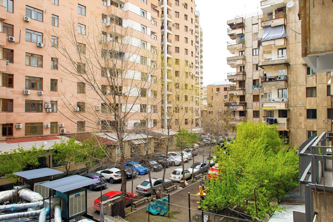 A multi-story residential building with balconies, air conditioning units, and a parking area with cars.