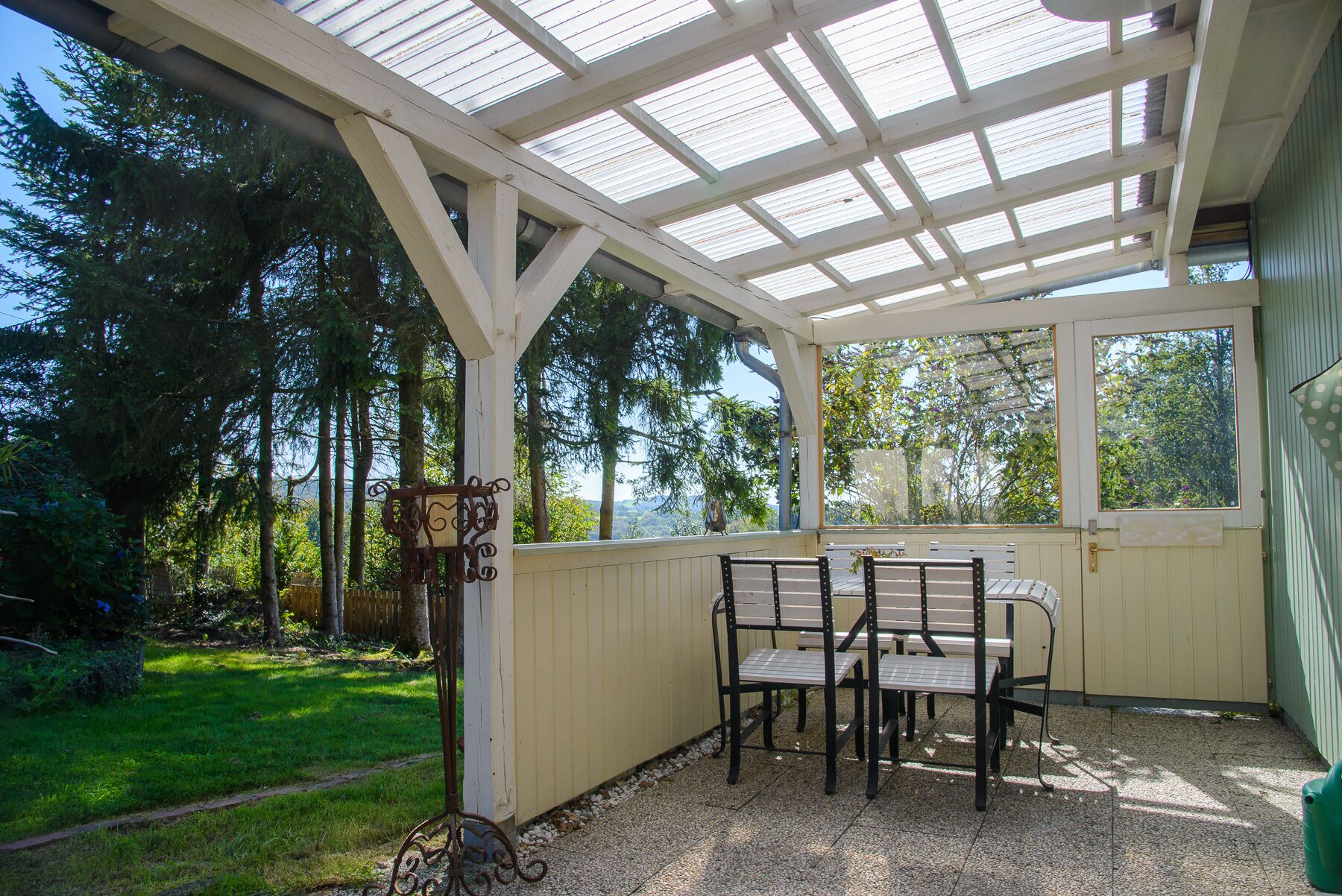 A covered patio with a table and chairs, surrounded by a garden and trees.
