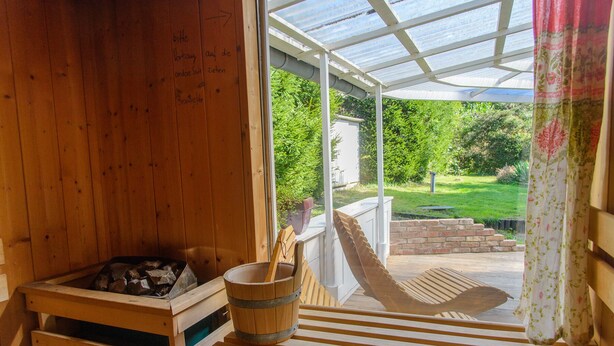 Una sauna in legno con una panca, un secchio, una porta in vetro che dà sull’esterno e vista sul giardino.
