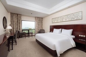 A hotel room with a large bed, a desk, a chair, a window with curtains, and a painting on the wall. - Nanyue Resort (Huoshan)