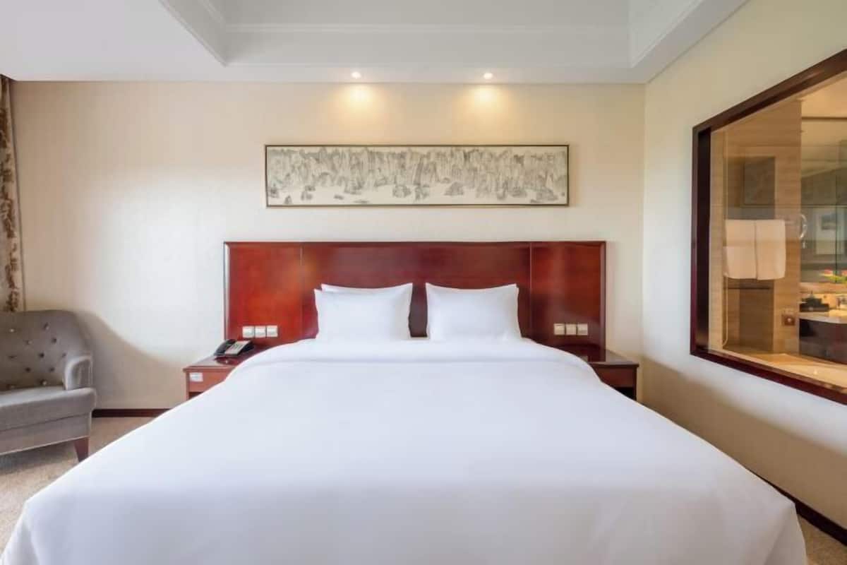 A hotel room with a large bed, a wooden headboard, a bedside table with a lamp, a gray armchair, and a large window.