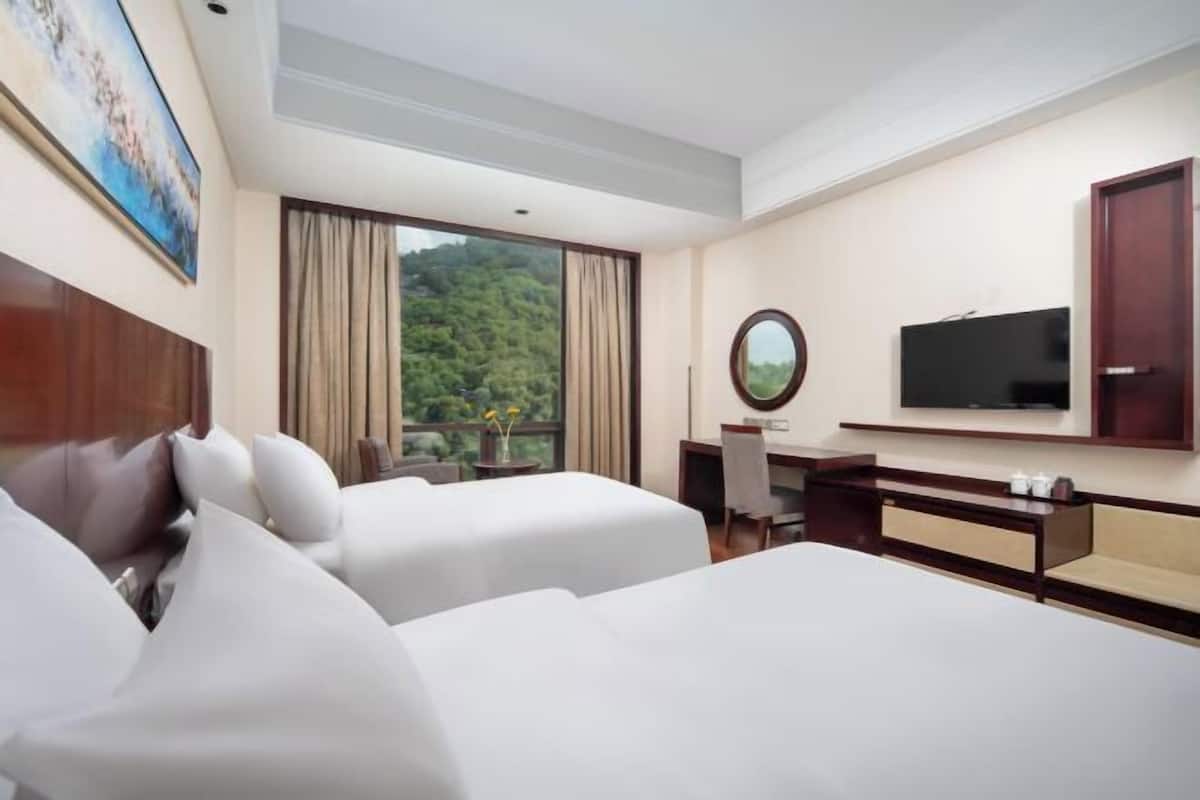 A hotel room with two beds, a desk, a TV, and a window with a view of greenery.