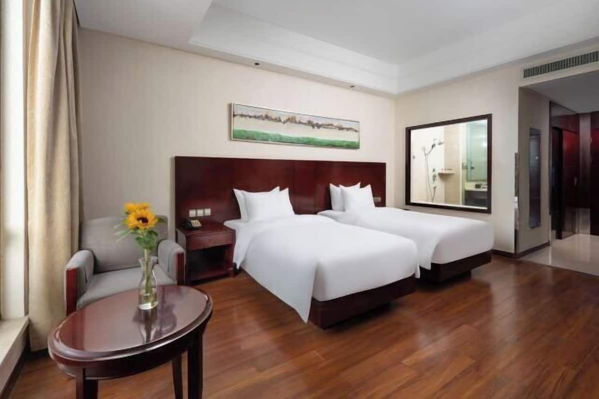A hotel room with two beds, a small table with a vase, a chair, a painting on the wall, and a mirror.