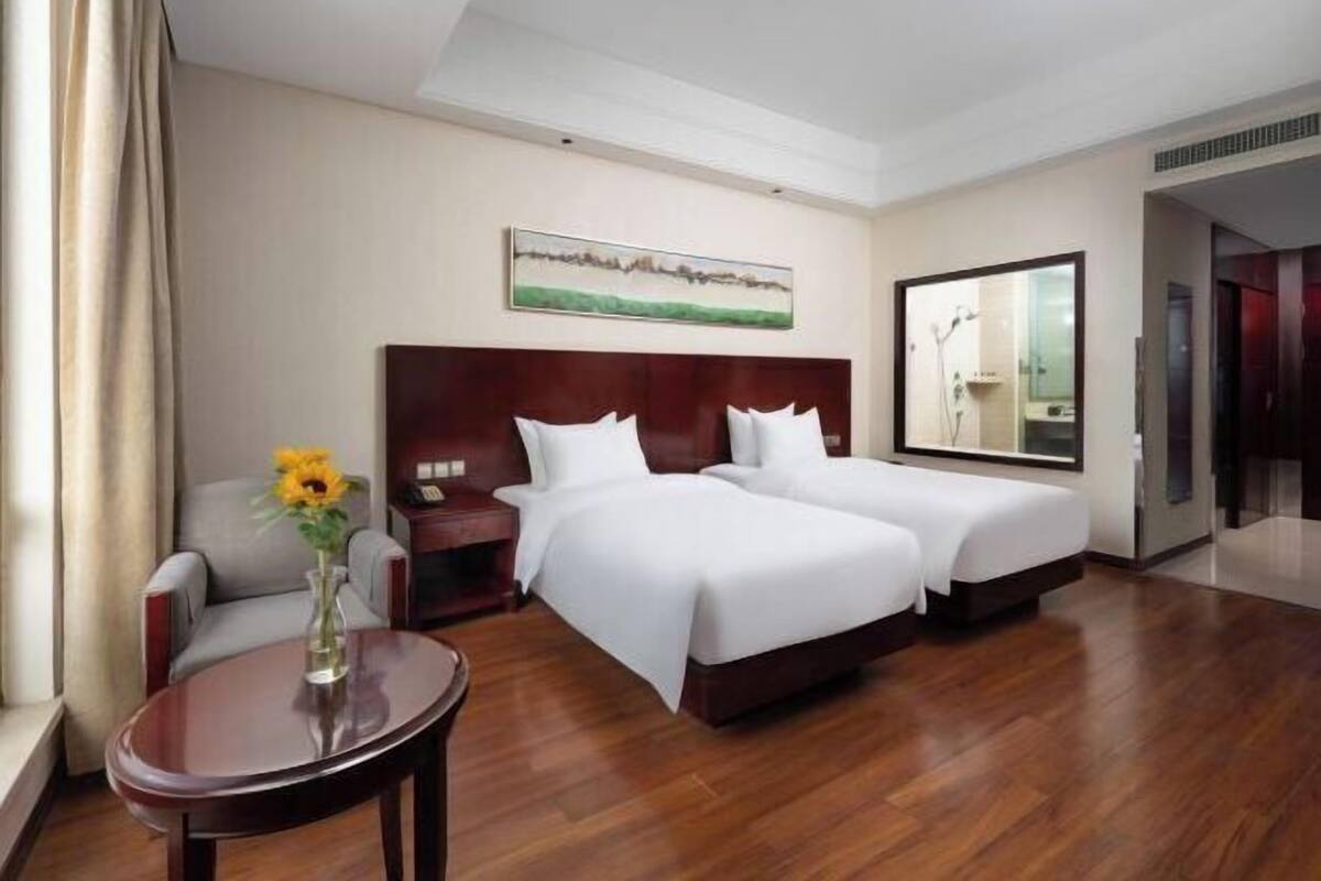 A hotel room with two beds, a small table with a vase, a chair, a painting on the wall, and a mirror.
