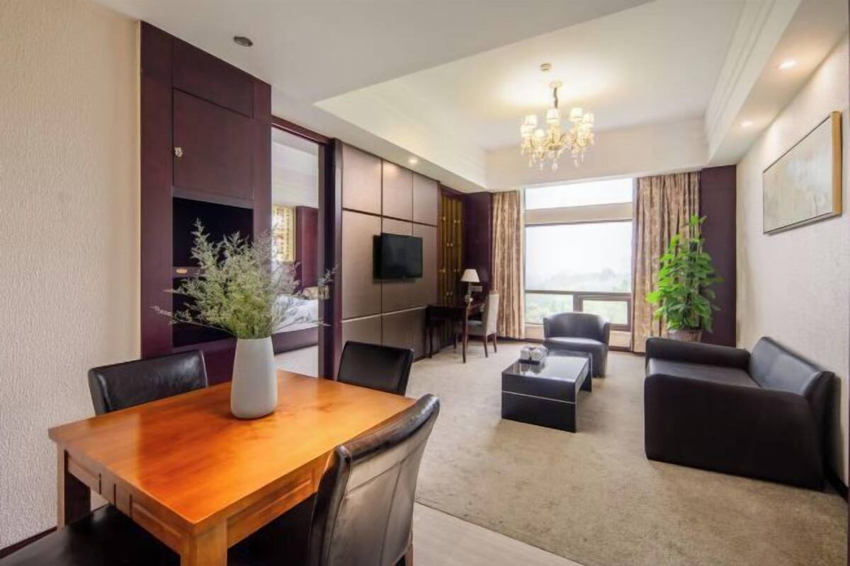 A hotel room with a wooden dining table, black leather chairs, a sofa, a TV, and a large window.