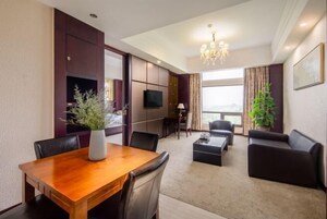 A modern hotel room with a wooden dining table, black leather chairs, a sofa, a TV, and a large window with curtains.