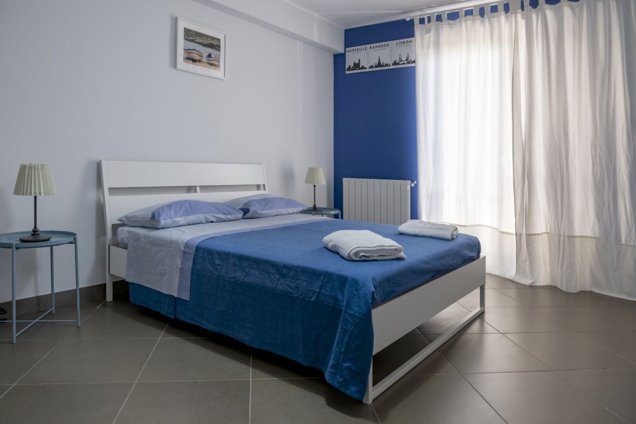 A bedroom with a bed, two bedside tables, a wall-mounted radiator, and a window with curtains.