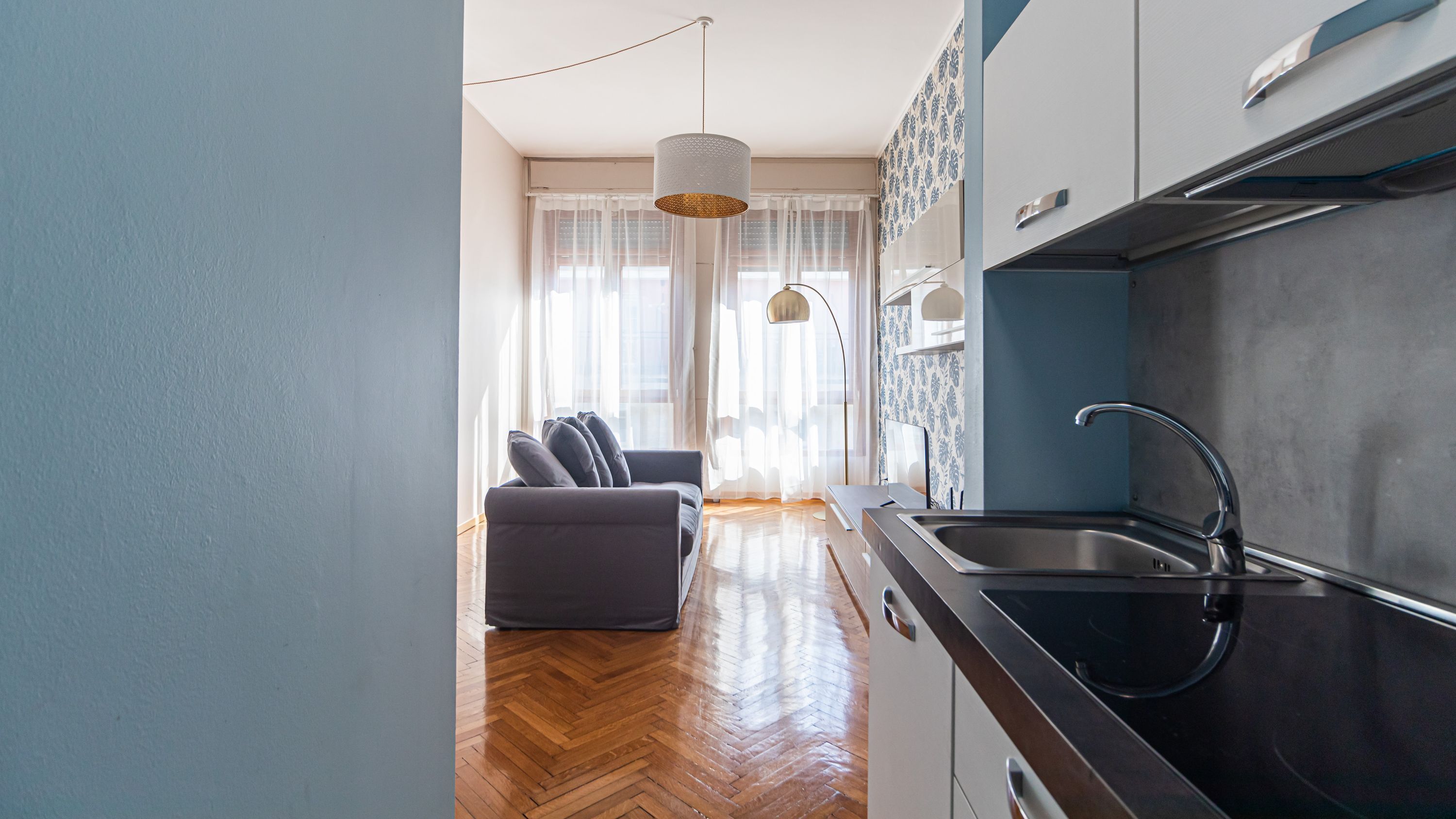 Una cocina moderna con encimera negra y pileta de acero inoxidable, sofá gris y un amplio ventanal con cortinas translúcidas.