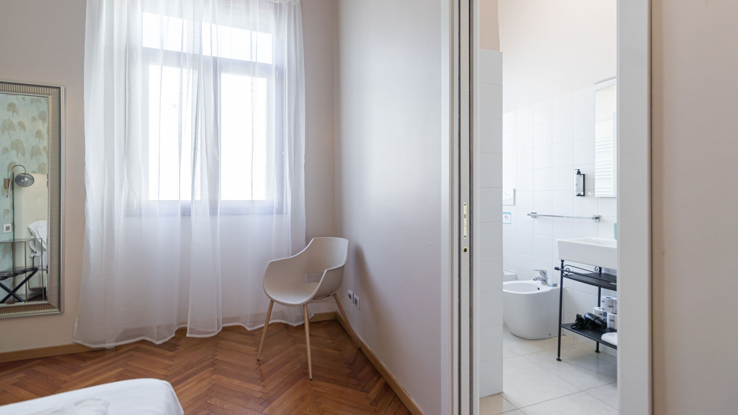 A hotel room with a bed, a chair, a window with sheer curtains, and a view into the bathroom.