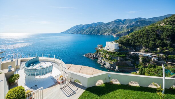 Ein Küstenhotel mit einer runden Außensitzgelegenheit, einem Pool und Blick auf das Meer und die Berge.