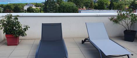 Zwei Liegestühle auf einer gefliesten Terrasse mit Blick auf Berge und ein Gewässer.