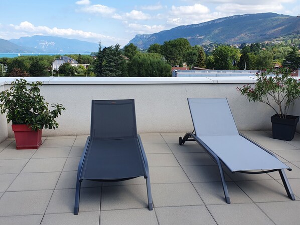 Two lounge chairs on a tiled terrace with a view of mountains and a body of water.