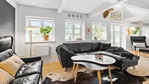 A modern living room with a black leather sectional sofa, a white coffee table, and a cowhide rug. There are large windows with white frames, a radiator, and a ceiling with exposed beams. - Stunning home in Nørre Nebel (Nørre Nebel)