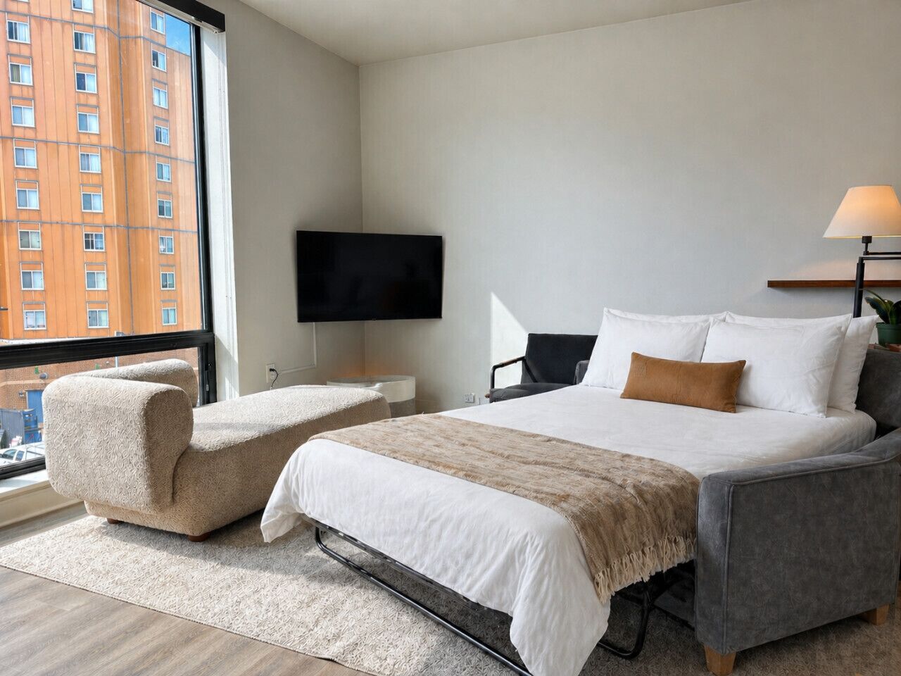A modern hotel room with a large bed, two armchairs, a flat-screen TV, and a view of a building through a large window.