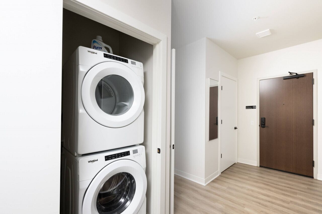 Une buanderie équipée de deux machines à laver et deux sèche-linge Whirlpool superposés, d’une porte en bois et d’un couloir avec un interrupteur et une poignée de porte.