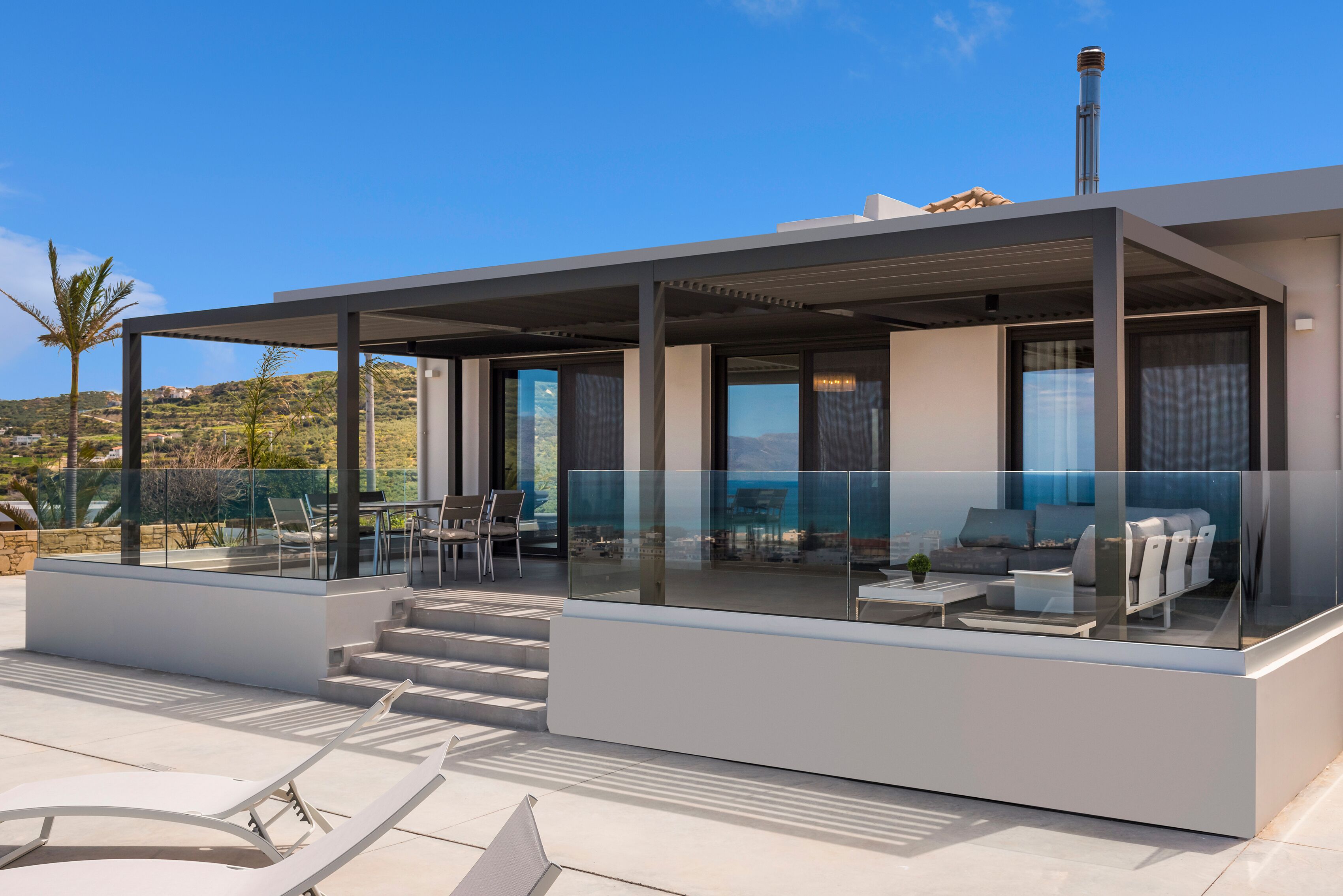 Ein modernes Haus mit einer glasvergrößerten Terrasse, Gartenmöbeln und Blick auf die Landschaft.