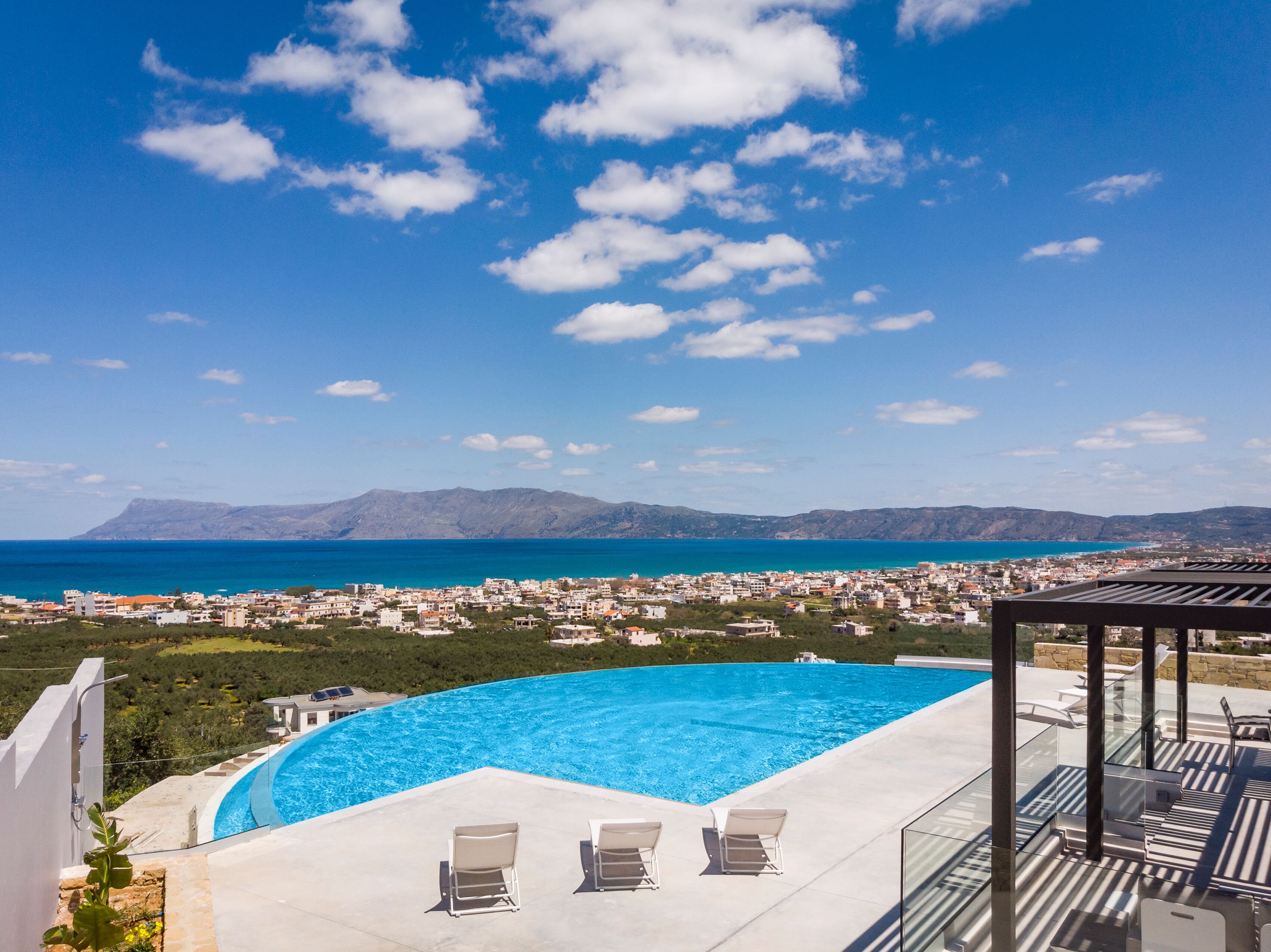 A swimming pool with lounge chairs, a clear view of the sea, and a mountainous landscape in the background.