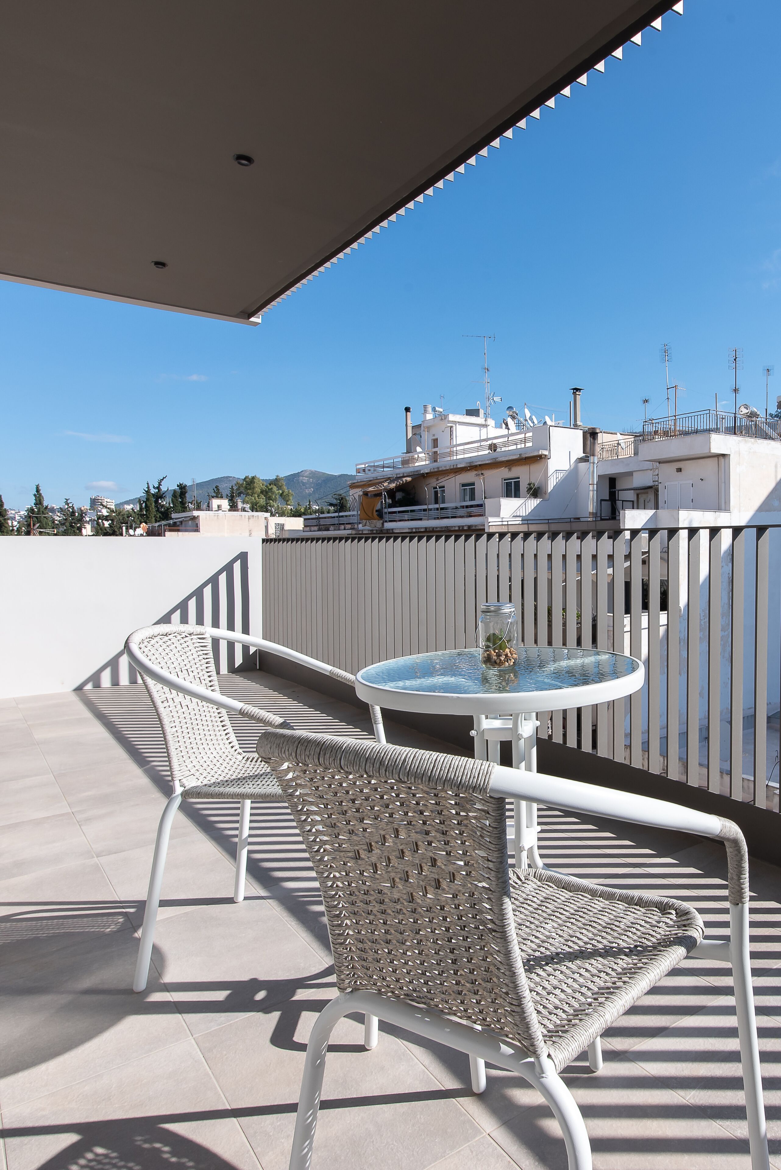 Un balcon avec une table en métal blanc et deux chaises en osier, donnant sur un paysage urbain avec des bâtiments et des montagnes.
