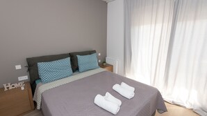 Een netjes opgemaakt bed met twee opgerolde handdoeken, een grijs gewatteerd sprei en een hoofdbord met gekleurde kussens.
