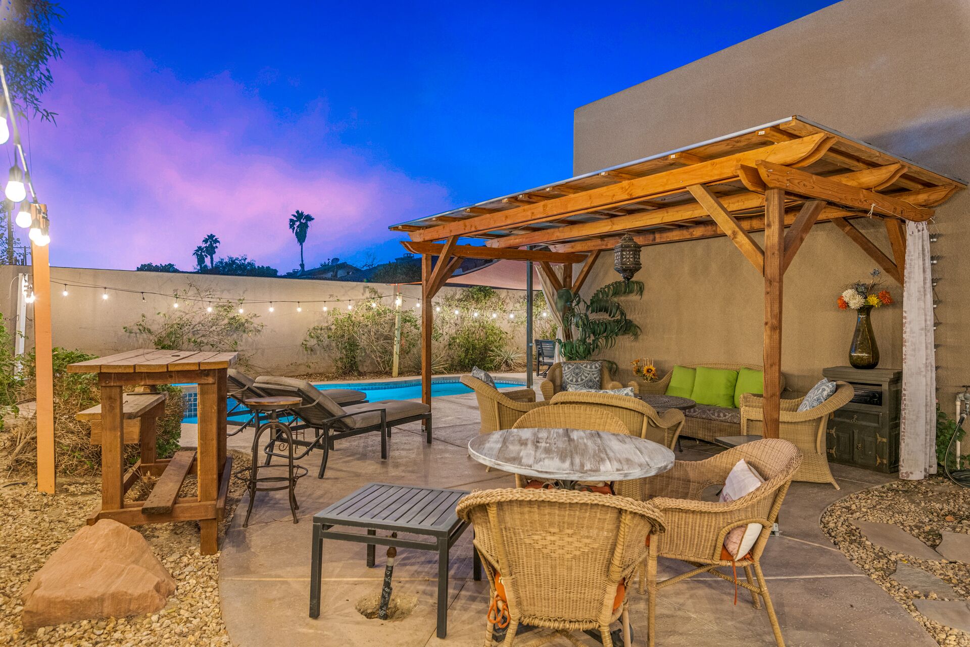 Patio esterno con piscina, pergola in legno, mobili in vimini e luci a stringa.