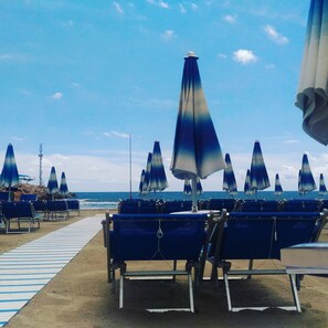 A beach with rows of blue beach chairs and umbrellas, a wooden walkway, and the ocean in the background.