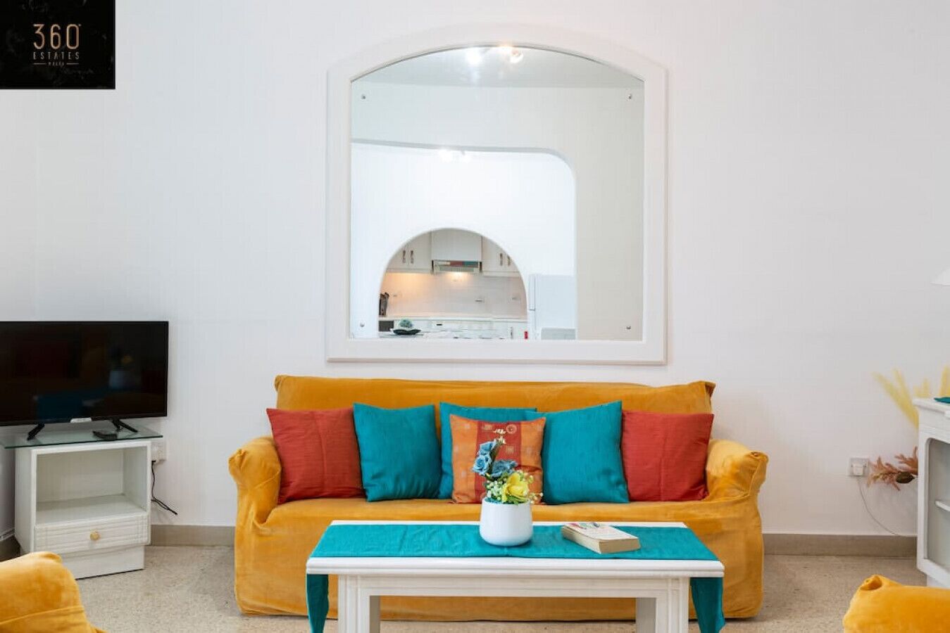 Ein Wohnzimmer mit gelbem Sofa, einem großen Spiegel, einem Fernseher auf einem Schrank und einem kleinen Tisch mit einer Pflanze.
