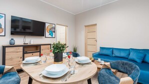 En moderne stue med et rundt spisebord dækket til fire personer, en blå sofa og et fladskærms-tv monteret på væggen.