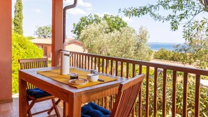 Une table en bois, sur laquelle sont posés un pichet blanc, un bol et une assiette remplie de nourriture, est placée sur un balcon offrant une vue sur la mer et la verdure.