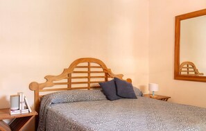 Un dormitorio con cabecera de madera, colcha estampada, almohada oscura, mesita de noche con lámpara y espejo en la pared.