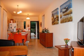 A modern living room with a sofa, a TV, and a wooden cabinet. There is a dining area with orange chairs and a kitchen in the background.