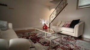 A living room with a white sofa, a glass coffee table with a red vase, and a staircase with a metal railing. - Momma Mary's Place @ Lawrenceville (Pittsburgh)