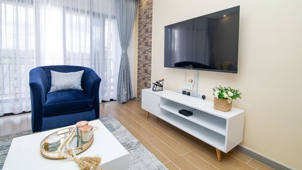A modern living room with a blue armchair, a white coffee table with decorative items, a flat-screen TV mounted on the wall, and a white entertainment unit. - Amani Suite by Homes By Della (Nairobi)