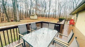 Une terrasse extérieure couverte, dotée d’une table à plateau de verre et de bancs, entourée d’une plateforme en bois et offrant une vue sur une zone boisée.