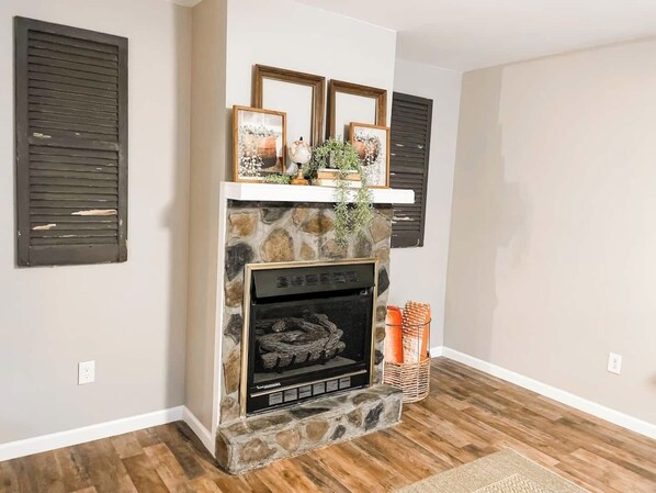 A fireplace with a stone surround, flanked by two dark shutters, and a mantel decorated with framed pictures and a plant.