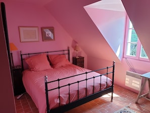 A bedroom with a sloped ceiling, a bed with pink bedding, two framed pictures on the wall, a nightstand with a lamp, and a window with a view of greenery.