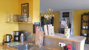Eine Küche mit Spüle, Kaffeemaschine und verschiedenen Küchenutensilien auf der Arbeitsfläche. Ein Bücherregal mit Büchern und eine Vase mit Blumen sind ebenfalls zu sehen.