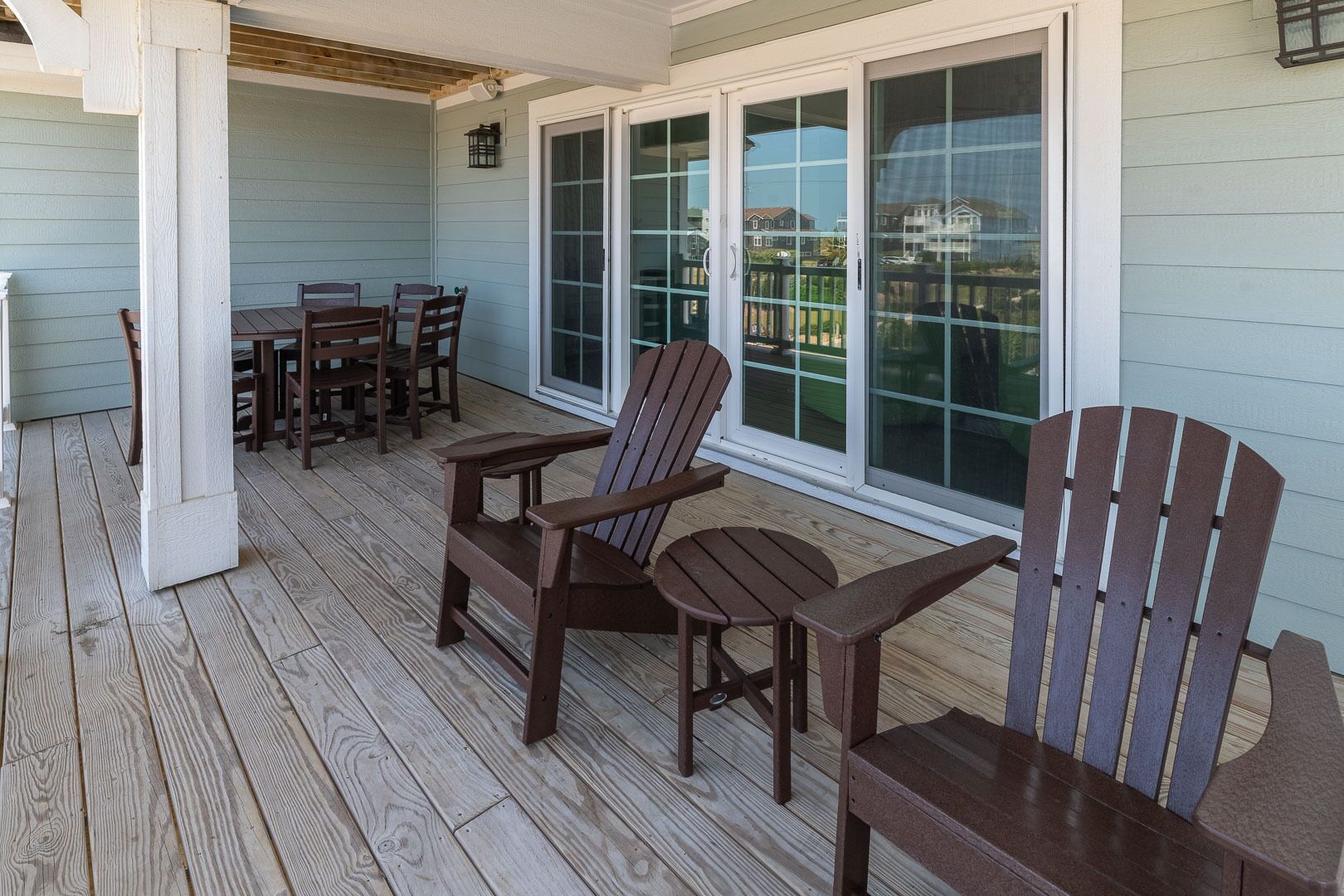 A covered porch with wooden chairs and a table, large windows, and a view of a residential area.