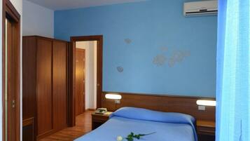 Quarto com piso de madeira, cama com colcha azul, mesa de cabeceira, aparelho de ar condicionado e porta.
